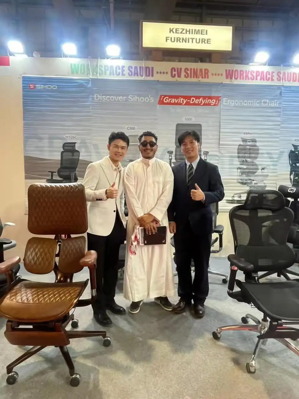 SIHOO personnel and Saudi customers take a group photo at the exhibition booth. All three smile and give a thumbs-up. The background is SIHOO's ergonomic chair display wall, highlighting the business cooperation and product display scene.