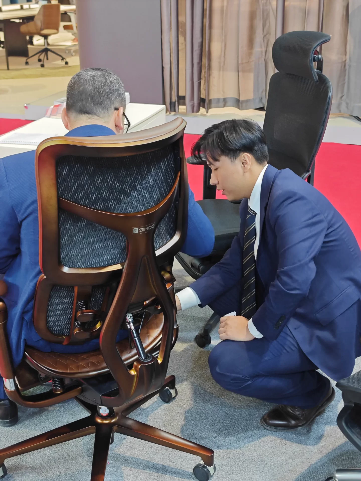 A SIHOO staff member explains the back adjustment details of an ergonomic chair to a customer at the Saudi exhibition. The customer experiences the chair, showing the chair's structural design and functional features.