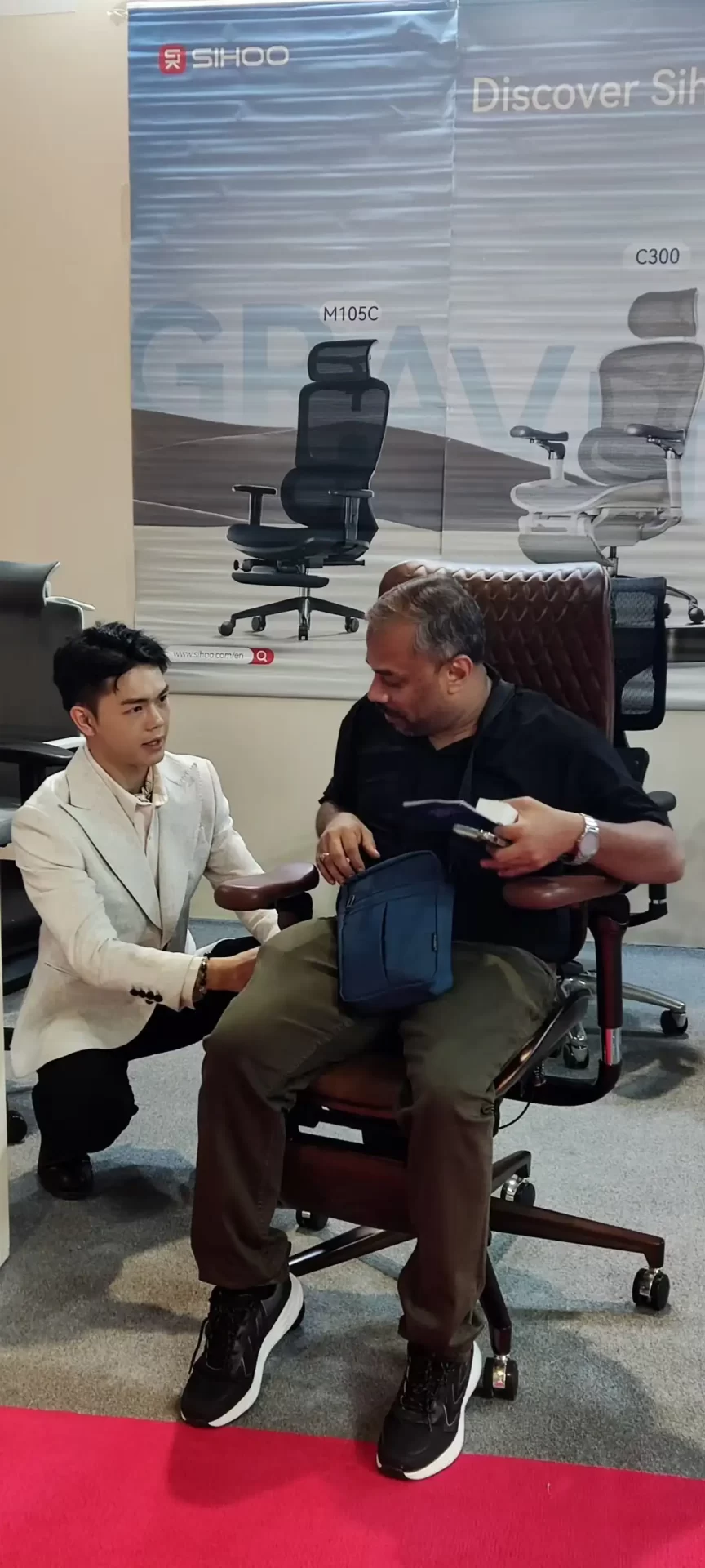 A SIHOO staff member explains an ergonomic chair to a customer at the Saudi exhibition. The customer experiences the chair, and the background wall displays models like M105C, presenting the product experience and explanation scene.